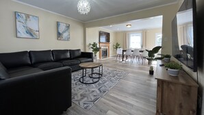 A modern living room with a black leather sofa, a coffee table, and a chandelier. There is a dining area with a table and chairs, and a TV mounted on the wall.
