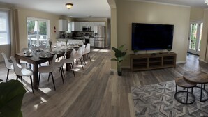A modern living room with a dining area, a large flat-screen TV, and a kitchen in the background.