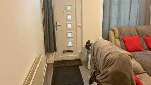 A room with a door, a radiator, a small rug, a sofa with red cushions, and a window with curtains.