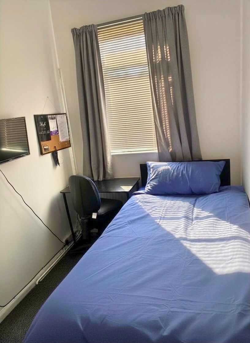 A single bed with a blue mattress and pillows, a desk with a chair, a window with curtains, and a bulletin board on the wall.