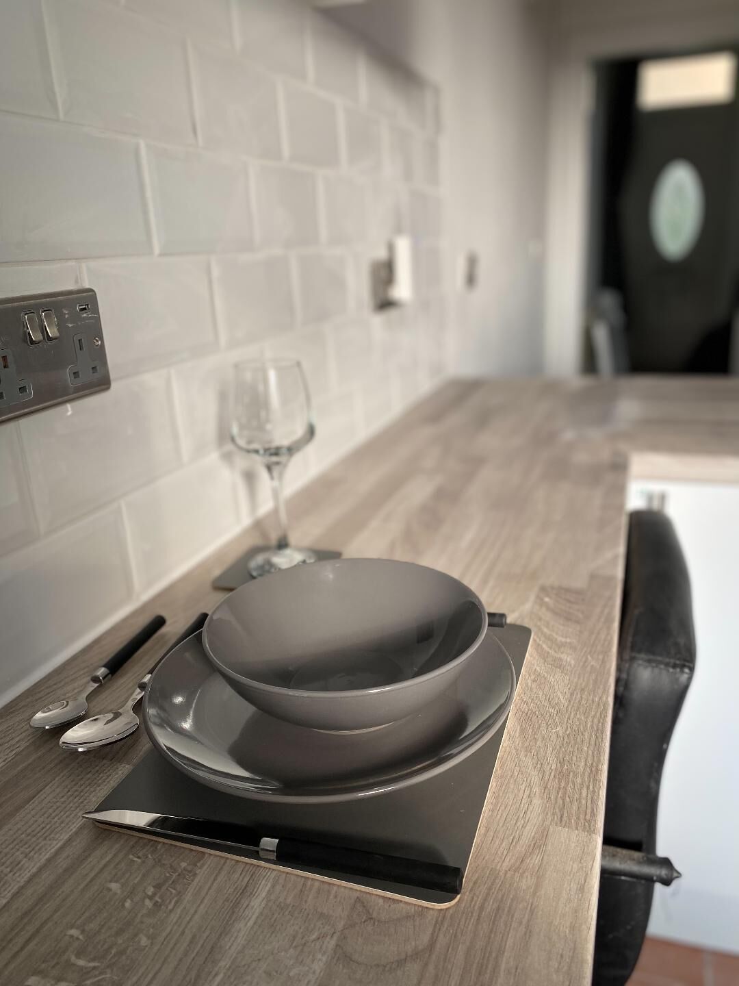 A modern kitchen with white tiled backsplash, a wooden countertop, a black pan on a cutting board, a wine glass, and a black chair.