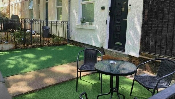 A residential building with a white exterior, a black door, and a small garden area with artificial grass and two chairs.