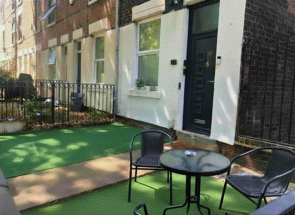 A residential building with a white exterior, a black door, and a small garden area with artificial grass and two chairs.