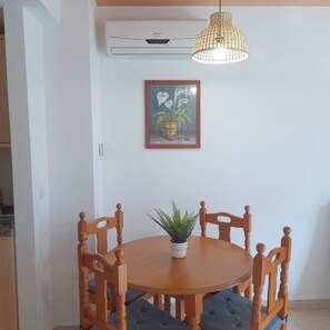 Un comedor con una mesa de madera, cuatro sillas, una planta en maceta, una lámpara colgante y un equipo de aire acondicionado en la pared.