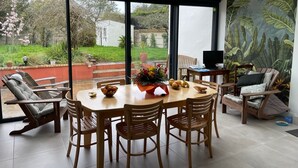 Ein Essbereich mit Tisch und Stühlen, ein Laptop auf einem Beistelltisch und Blick auf einen Garten durch große Fenster.