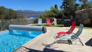 A swimming pool with a view of mountains, a barbecue grill, and two lounge chairs.
