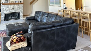 A spacious living room with a black leather sectional sofa, a fireplace, and a flat-screen TV mounted on the wall. - Condo in world class Leelanau Peninsula, Suttons Bay Beach Condo (Suttons Bay)