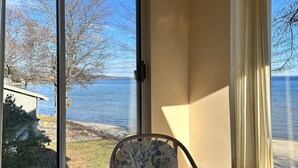 A room with a glass table and a chair, large windows with a view of a body of water and trees, and a curtain on one window. - Condo in world class Leelanau Peninsula, Suttons Bay Beach Condo (Suttons Bay)