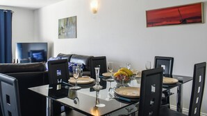 A dining area with a glass table set for four, surrounded by black chairs. There are two paintings on the walls, one abstract and one with a red background. A flat-screen TV is mounted on the wall to the left. - Apartment 15 Harvest Moon (Newquay)