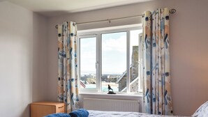 A bedroom with a bed, a window with curtains, a radiator, and a view of a coastal area. - Apartment 15 Harvest Moon (Newquay)