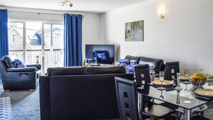 A modern living room with a sofa, armchairs, a flat-screen TV, and a dining table set for four. - Apartment 15 Harvest Moon (Newquay)