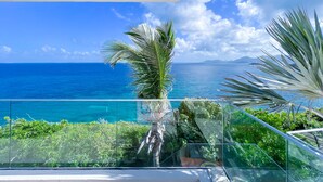 Ein Balkon mit Glasgeländer, der Blick auf ein klares, blaues Meer und ferne Berge gewährt.