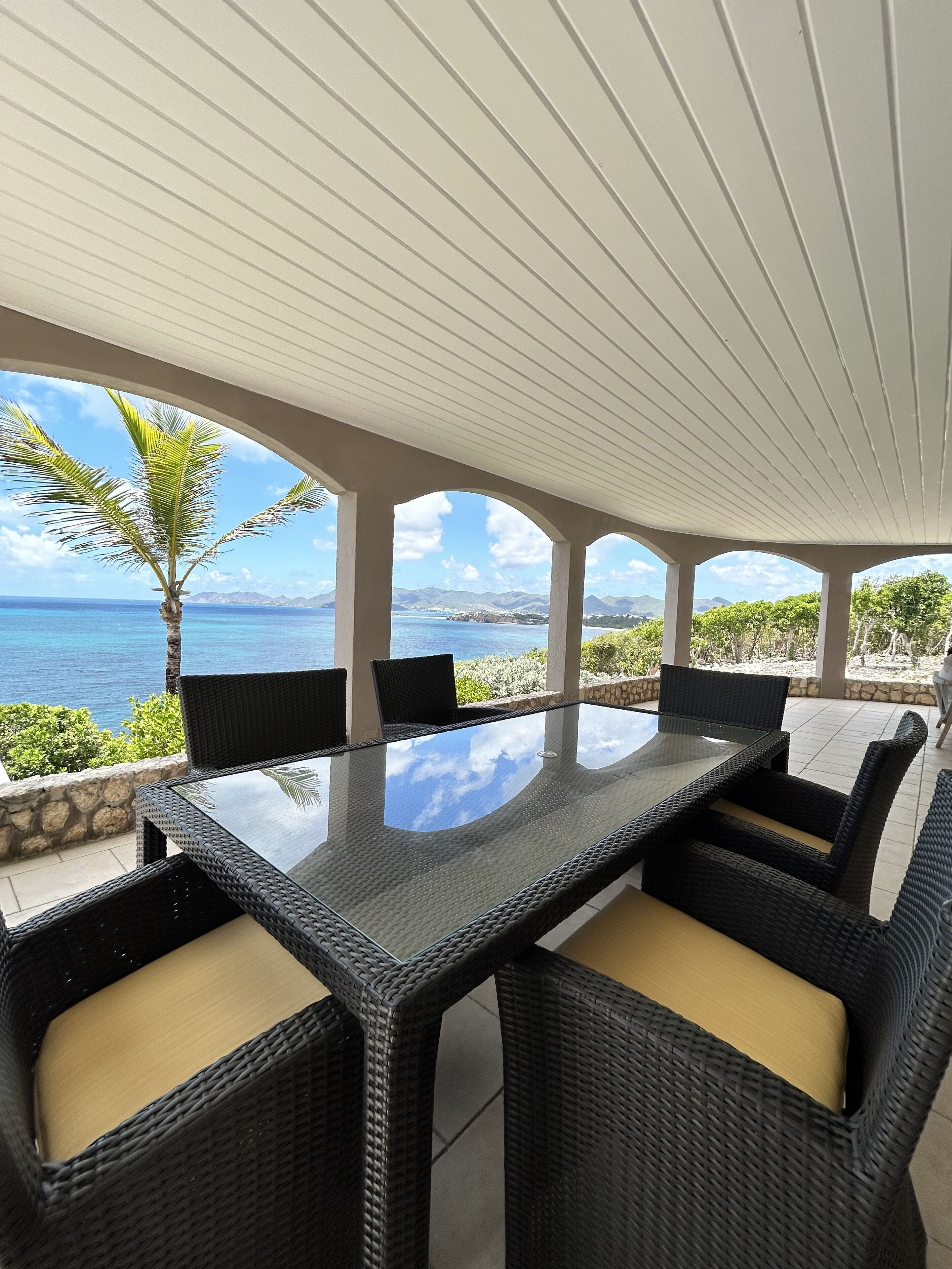An outdoor dining area with a glass-top table and wicker chairs, overlooking a scenic ocean view.