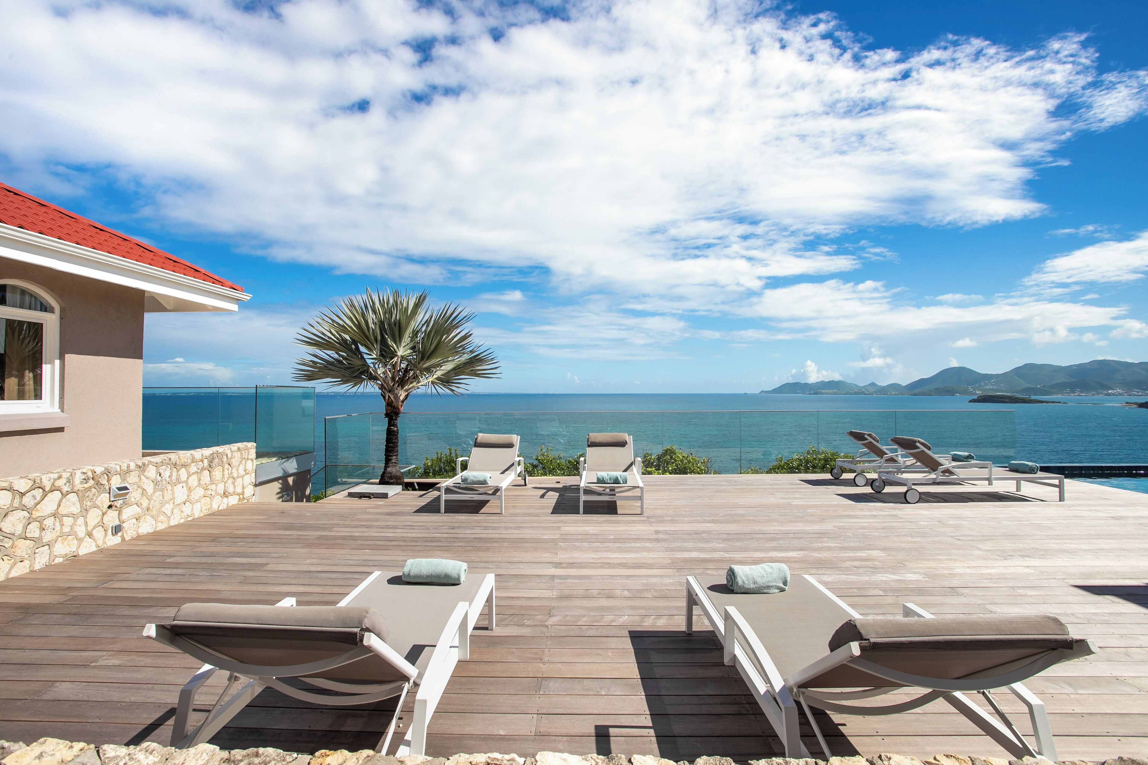 A wooden deck with lounge chairs and a palm tree, overlooking a body of water and distant mountains.