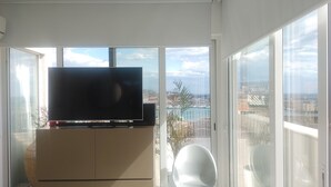 A balcony with a TV on a cabinet, large windows with a city view, and a white chair. - PENTHOUSE ON CATANIA HARBOR AND VOLCANO VIEW A FEW STEPS FROM CATHEDRAL (Catania)