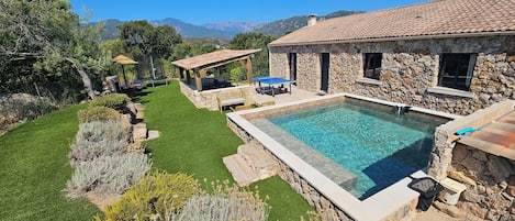 A stone building with a swimming pool, outdoor seating, and a view of mountains.