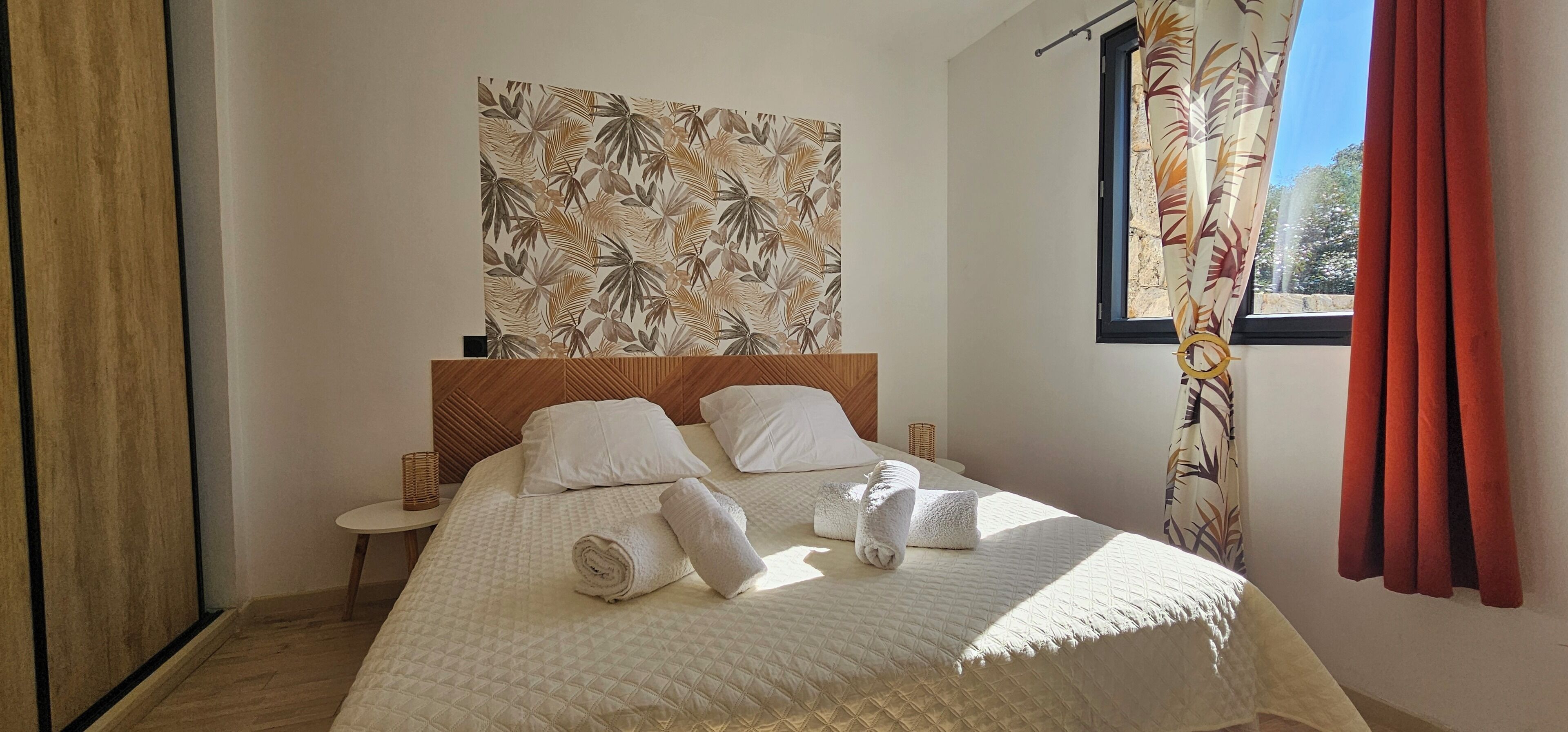 A bedroom with a bed, pillows, a headboard, a wall tapestry, a window with curtains, and a side table.