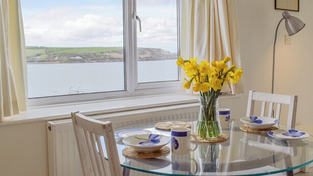 Un coin repas avec une table en verre, des chaises blanches et une vue sur une étendue d’eau, visible à travers une fenêtre.