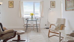 Un salon lumineux avec une table en verre, des chaises blanches, un canapé et une vue dégagée sur l’extérieur grâce à une grande fenêtre.