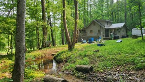 Une cabane dotée d’une terrasse et de meubles de jardin, entourée d’une forêt et bordée d’un petit ruisseau.