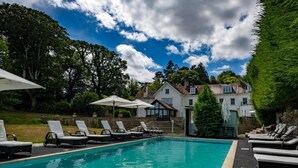 Una piscina con sillas de descanso y sombrillas, rodeada de árboles y un edificio al fondo.