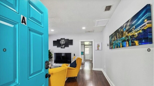 A hallway with a turquoise door, a yellow armchair, a flat-screen TV, and a cityscape painting on the wall. - Charming Seminole Heights Stay with Free Parking (Tampa)