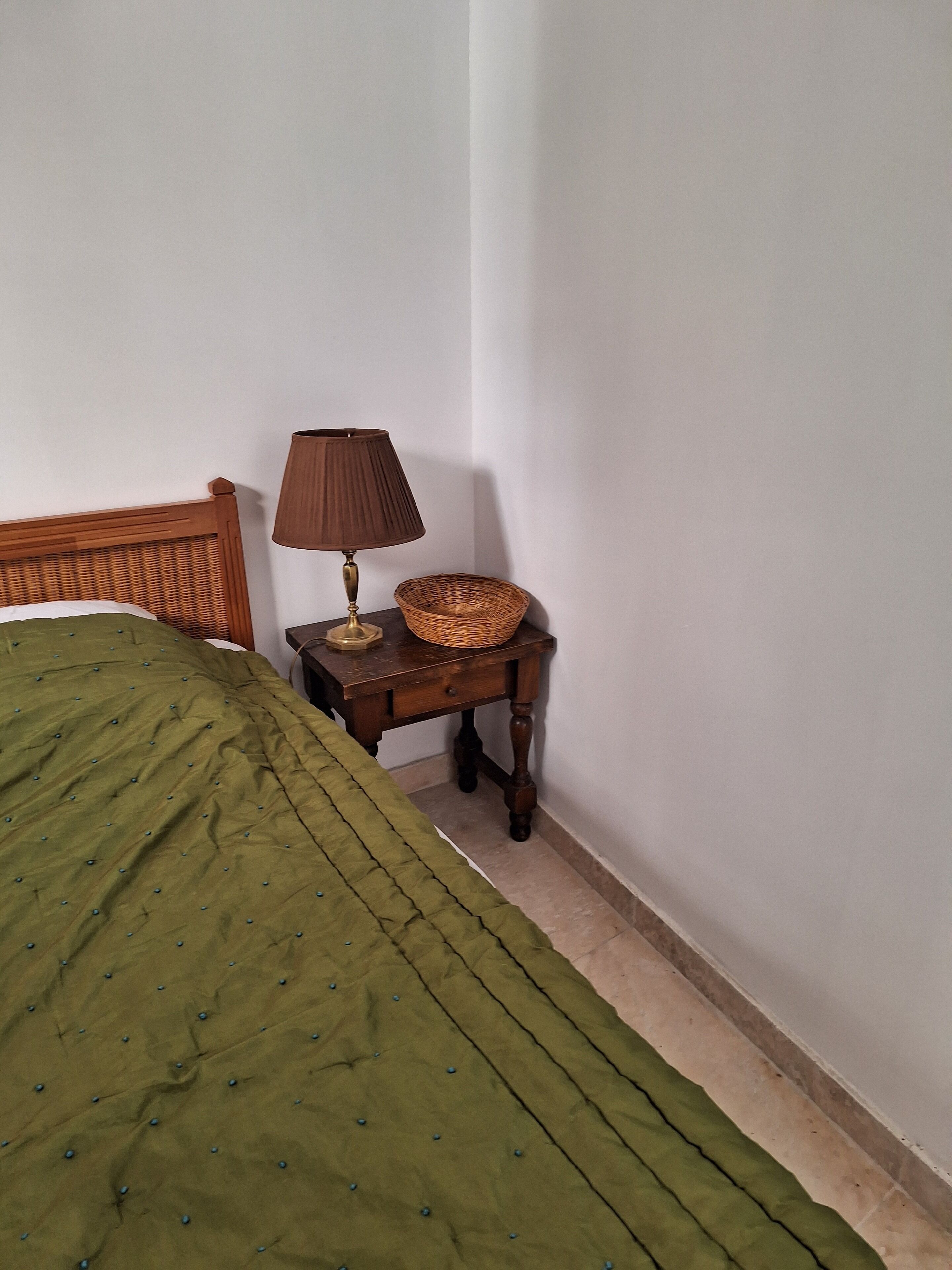 Un dormitorio con una colcha verde, una mesita de noche de madera con una lámpara y una cesta de mimbre.