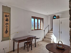 A room with a desk, chair, and a small table with a basket. There's a window with a view of mountains and a staircase leading to another level.