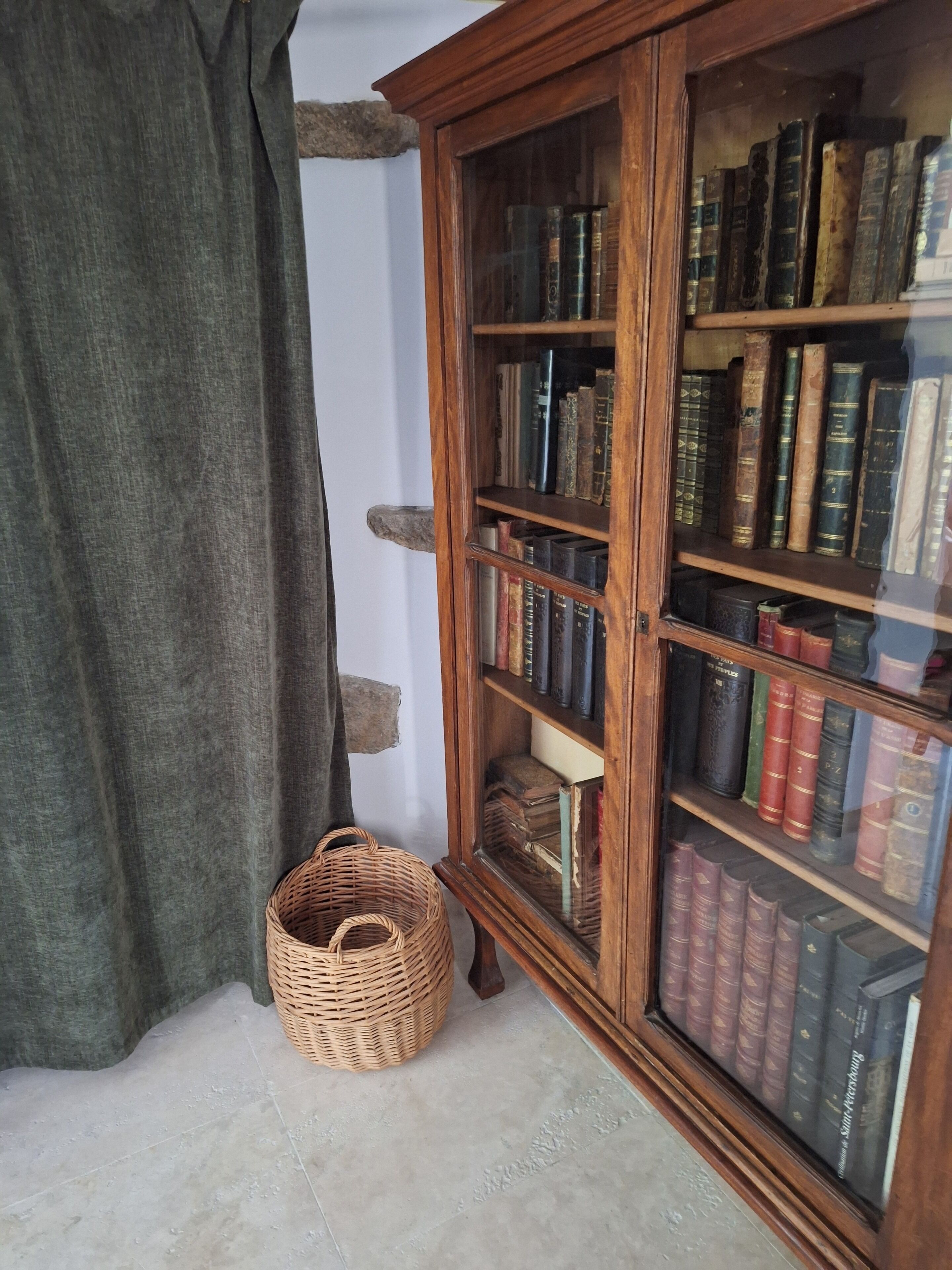 A wooden bookcase with glass doors filled with books, a wicker basket on the floor, and a curtain partially covering a window.
