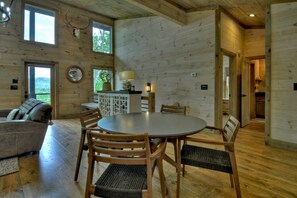 A wooden interior with a round dining table, chairs, a sofa, and a kitchen area.