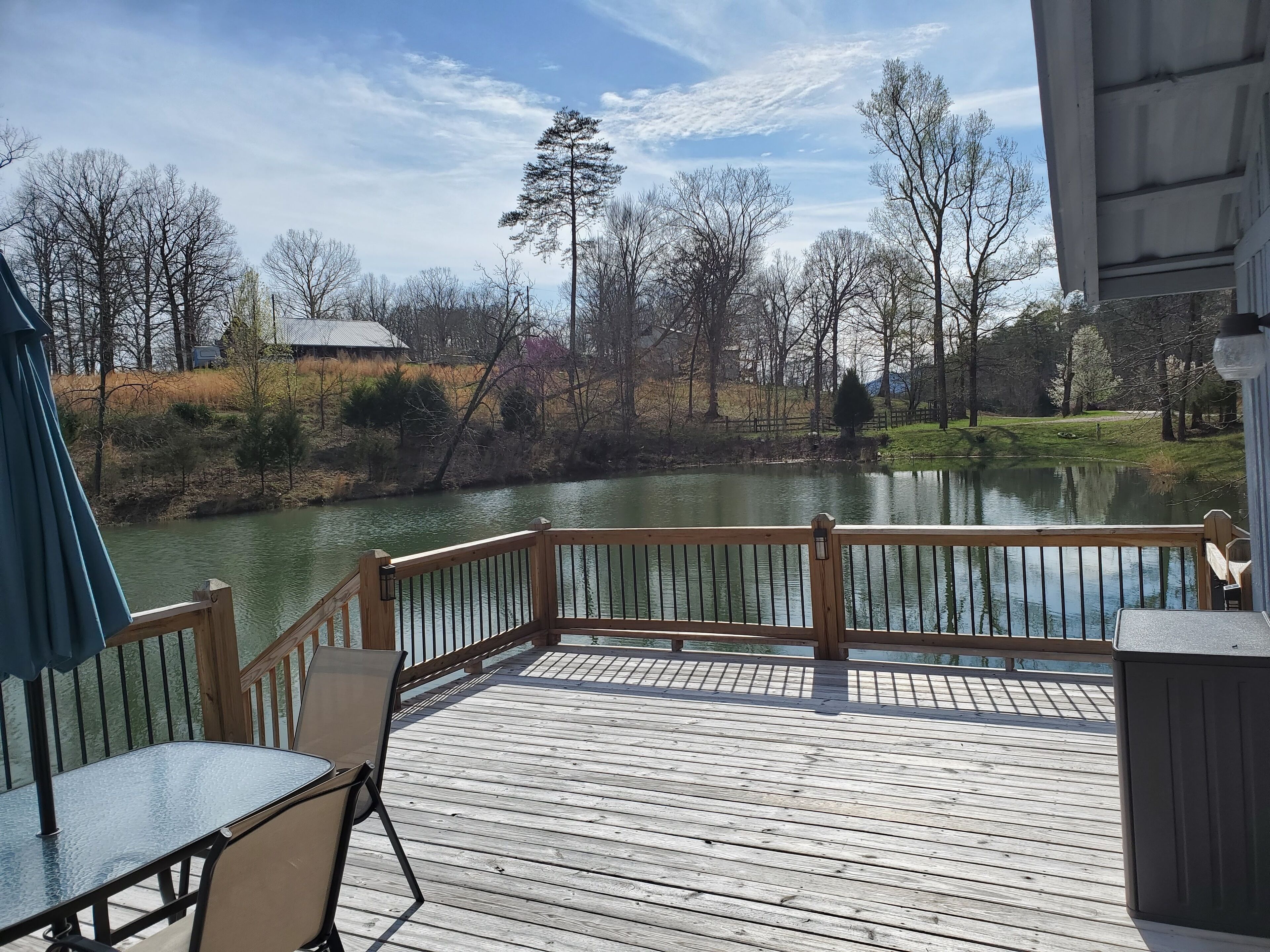 A wooden deck with a table and chairs, overlooking a calm lake with trees and a distant building.