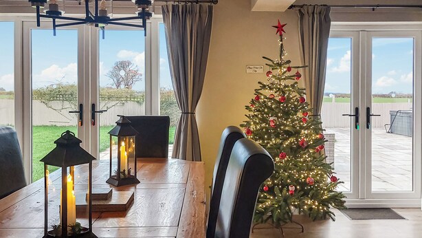 Uma sala de jantar com uma mesa de madeira, duas cadeiras pretas, um pinheiro de Natal com luzes e enfeites, e um candeeiro aceso.