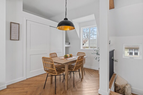 Een eetgedeelte met een houten tafel en rieten stoelen, een zwarte hanglamp, en een witte deur op de achtergrond.