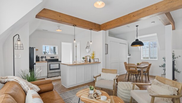 Un loft espacioso con techo alto, vigas de madera a la vista, una cocina con armarios blancos, un comedor con sillas de madera y una sala de estar con sofá de cuero marrón y una mesa de centro.