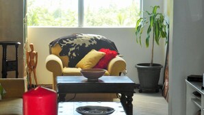 Une pièce avec une grande fenêtre, un canapé recouvert d’un plaid à motifs, une petite table sur laquelle se trouve un bol, une plante en pot et une bougie rouge.