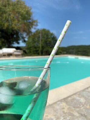 Un vaso con pajita de lunares y cubitos de hielo, situado junto a una piscina con cielo azul despejado y árboles al fondo.