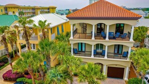 A multi-story building with a red-tiled roof, balconies with blue chairs, and a driveway with a car. - Suzie Q, Luxury Beach House w/Private Pool Rooftop Deck (Destin)