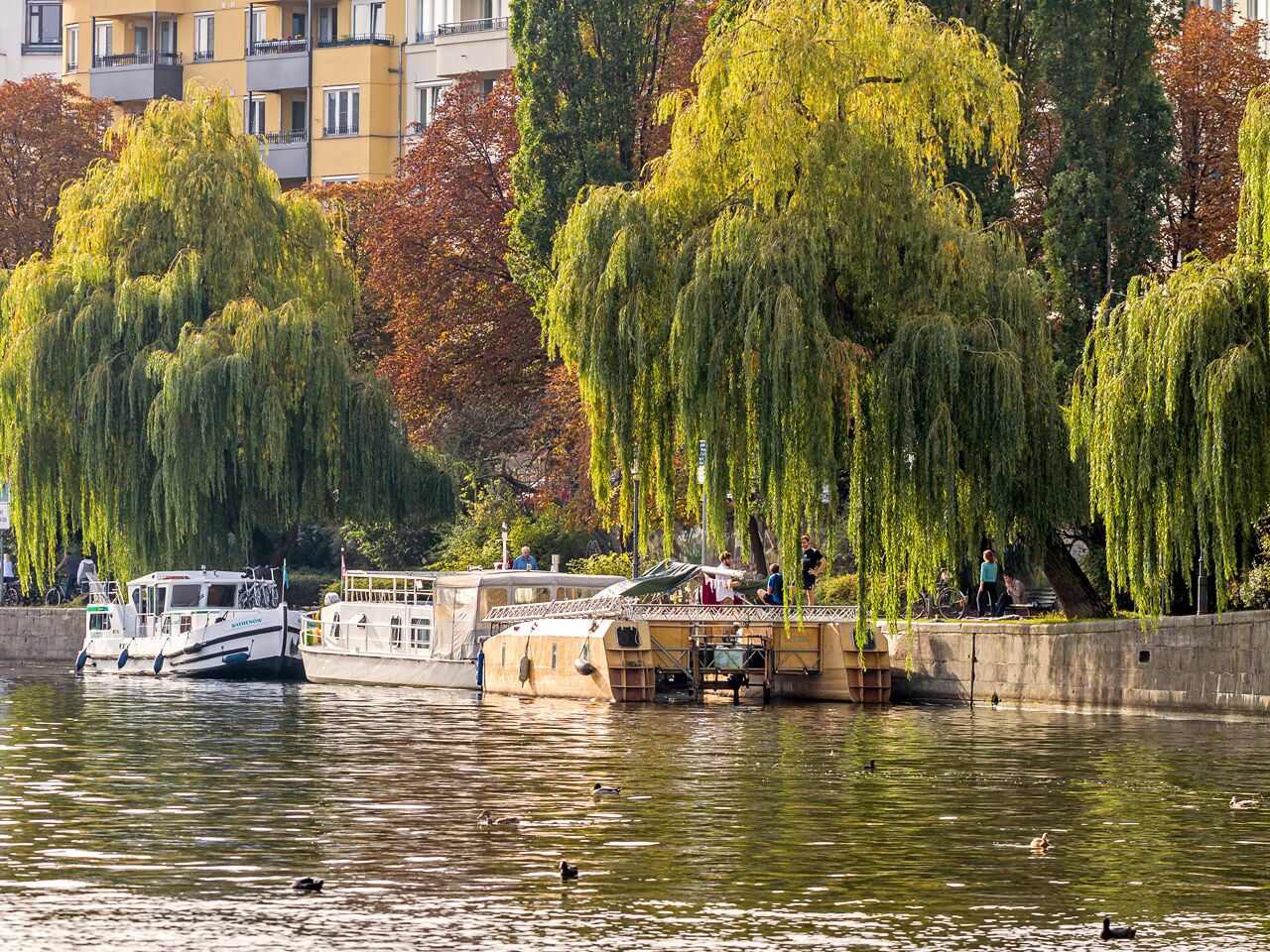 A river with boats docked, a pedestrian bridge, and trees with autumn foliage.