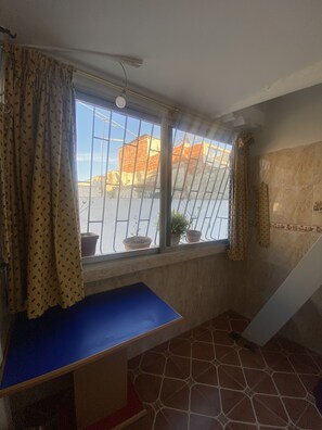 Ein Zimmer mit Fenster, einem blauen Sitzbänklein und Fliesenboden.