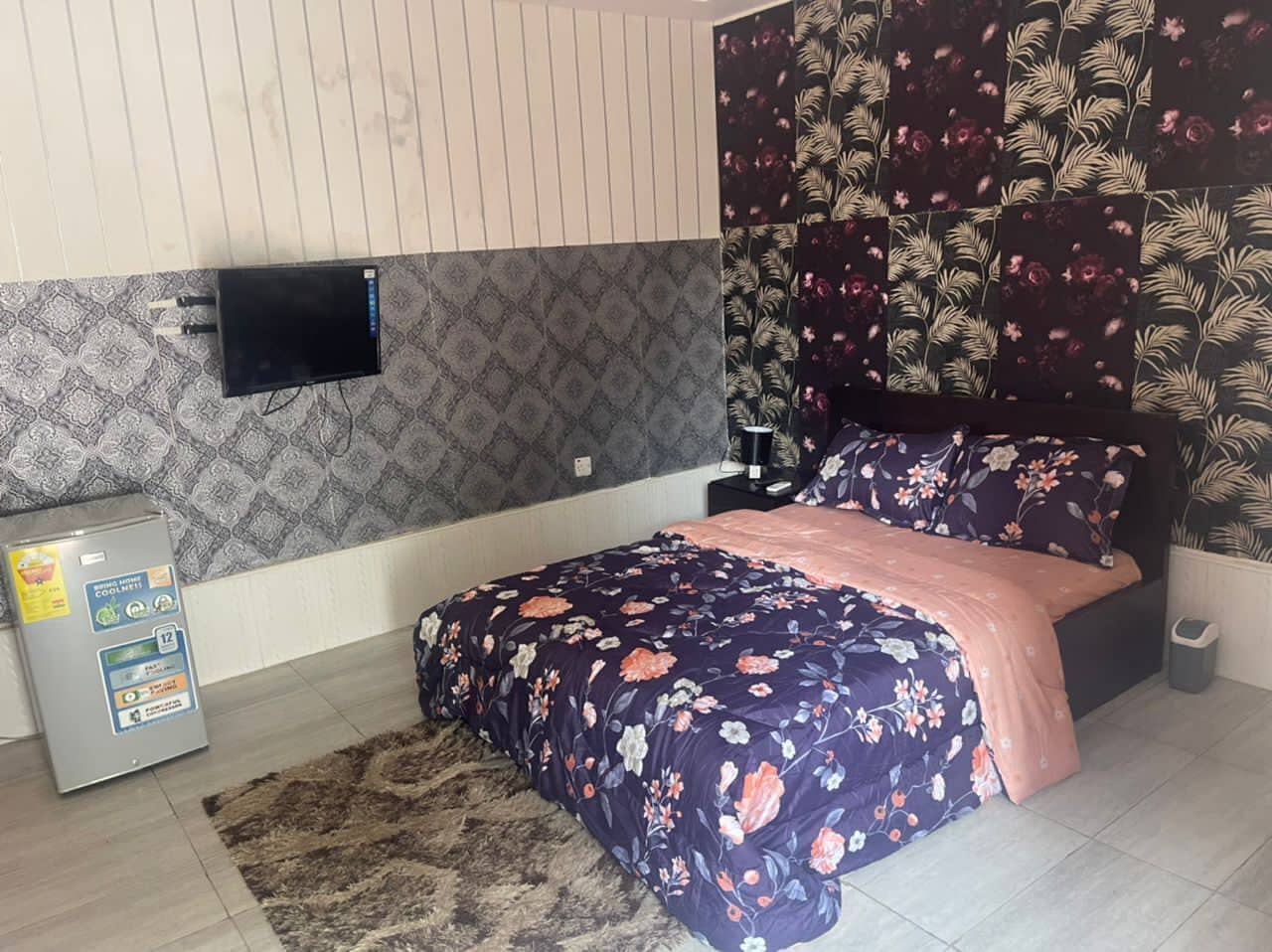 Un dormitorio con cama, televisión montada en la pared y un refrigerador pequeño.