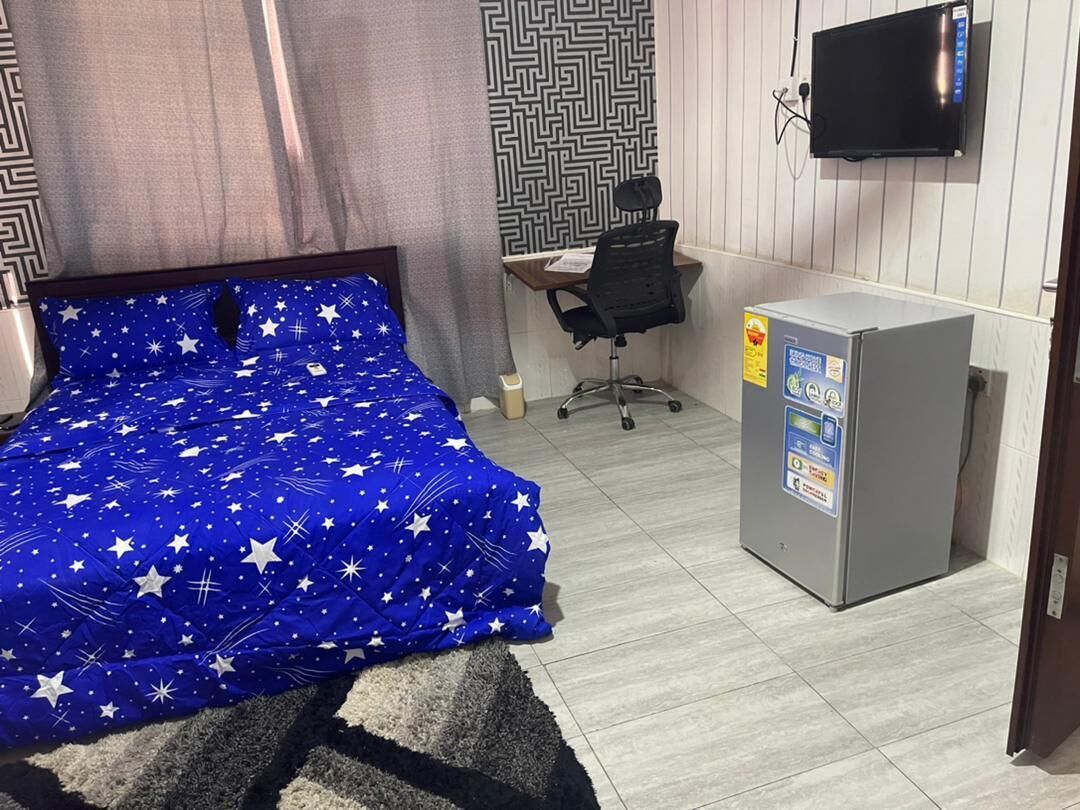 Une chambre avec un lit recouvert d’une couverture bleue à motifs d’étoiles, un petit bureau avec une chaise, un téléviseur fixé au mur et un réfrigérateur.