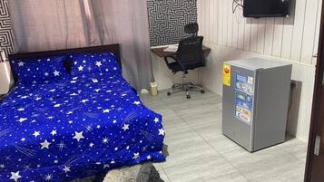 Une chambre avec un lit recouvert d’une couverture bleue à motifs d’étoiles, un petit bureau avec une chaise, un téléviseur fixé au mur et un réfrigérateur.