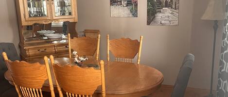 Une salle à manger avec une table ronde, six chaises en bois, un buffet en bois doté de portes en verre, un ventilateur de plafond et deux tableaux encadrés accrochés au mur.
