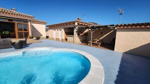 Une piscine avec une terrasse carrelée, un espace patio aménagé avec du mobilier d’extérieur, et un bâtiment doté d’un toit en tuiles et de murs en briques.