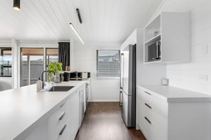 A modern kitchen with white cabinetry, a stainless steel refrigerator, and a window with blinds. - Be Rude Not To - Walk to Redwoods, Sleeps 7 (Rotorua)