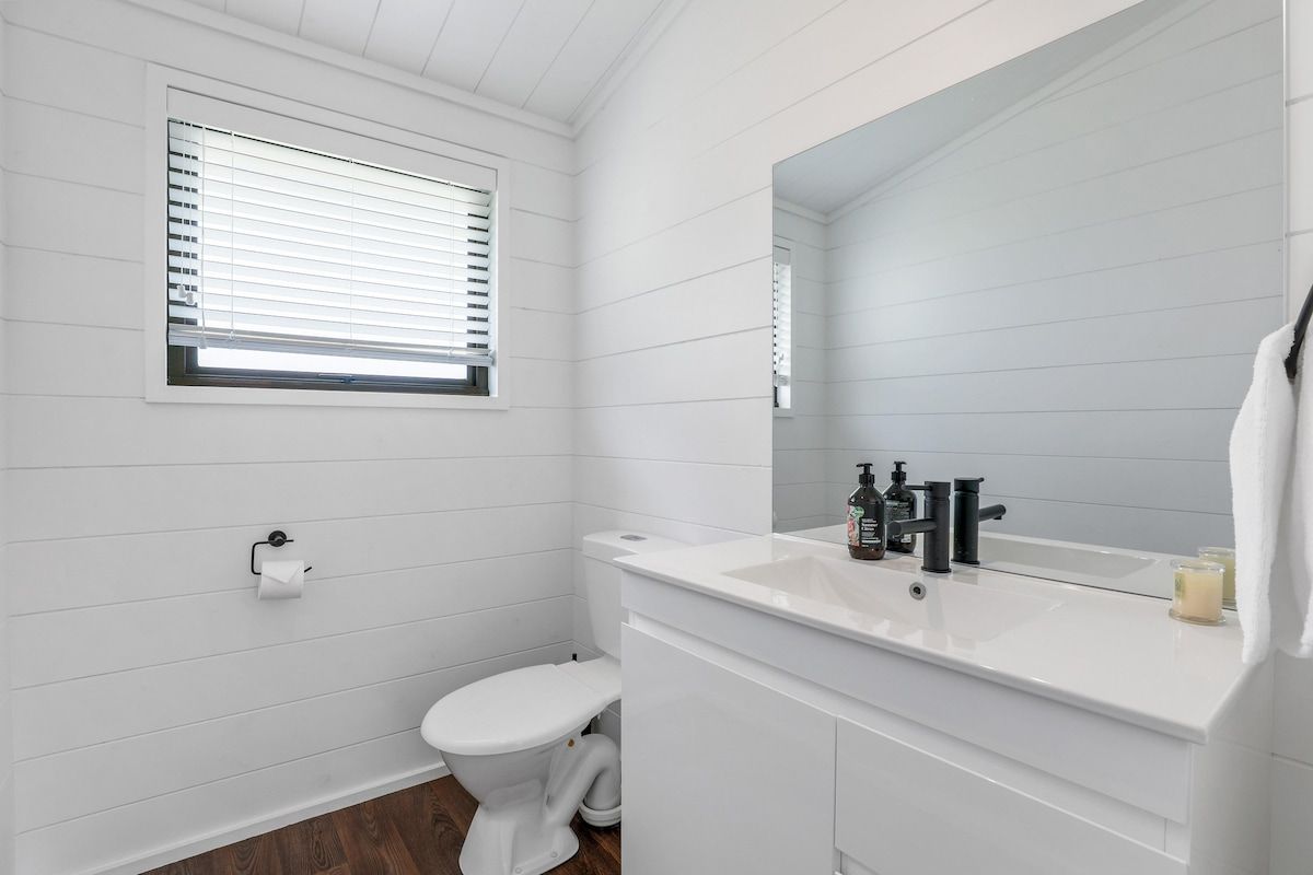Une salle de bain équipée de toilettes, d’un lavabo, d’un miroir et d’une fenêtre avec des stores.