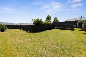 A fenced backyard with a grassy lawn, a small tree, and a building with a sloped roof. - Be Rude Not To - Walk to Redwoods, Sleeps 7 (Rotorua)