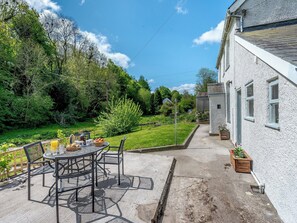 A patio with a round table and chairs, a small garden, and a white building with windows. - 2 bedroom accommodation in Knighton (Knighton)