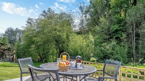 A round metal table with four chairs, surrounded by a wooden fence, set in a lush green garden with dense trees in the background.