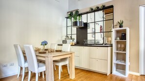 Ett modernt kök med vita köksskåp, ett matbord i trä och stolar. Köket har en inbyggd spis, ett upphängt hyllsystem för kastruller och en kaffemaskin på bänkskivan.